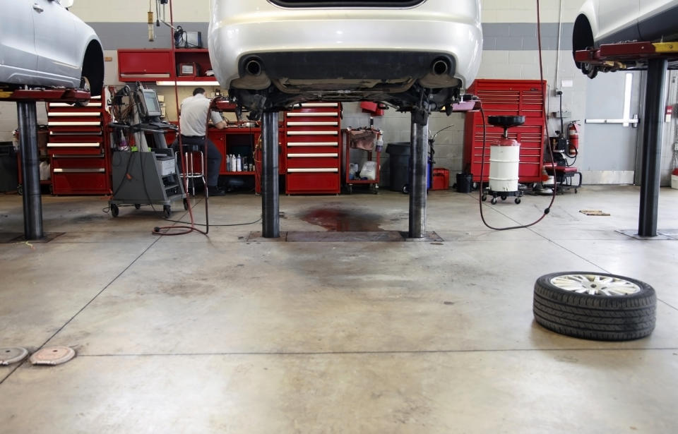 Car lifted on hydraulic jacks in an auto repair shop with red tool cabinets and a tire on the floor. Mechanics are visible in the background working