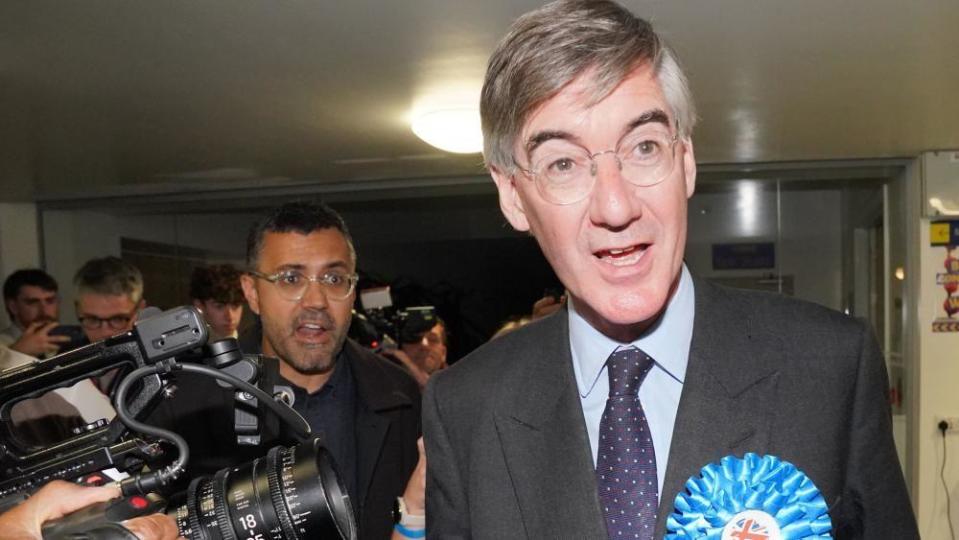 Jacob Rees-Mogg speaking with a camera next to him and a crowd behind