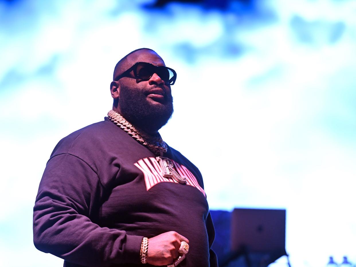 Rick Ross wearing black shades a black sweater and gold chains while standing on a brightly lit stage