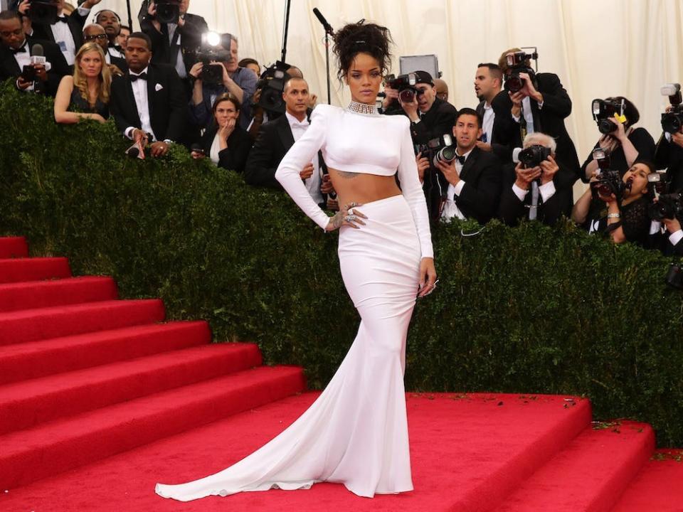 Rihannah poses on the Met Gala stairs wearing a cropped white top and flowing white skirt.