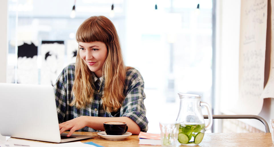 A woman sits at a cafe, with a coffee, looking at laptop.