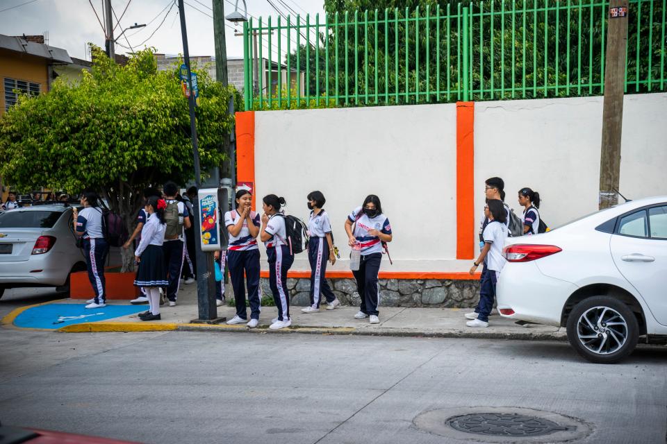 Students in uniform stand in front of a white structure on a busy street.