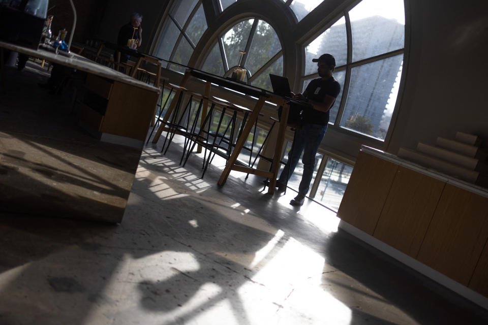 A person stands while looking at a laptop in front of a window in a shadowy room.