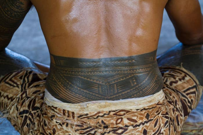 A person's back with traditional Polynesian tattoos is shown, accompanied by cloth with traditional patterns wrapped around the waist