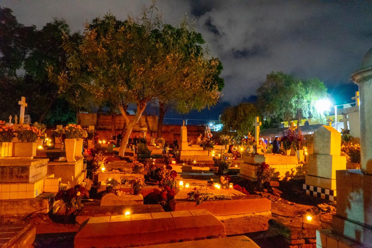 Xoxocotlán Cemetery, Oaxaca, Mexico, several candles lit up among the graves during the day of the dead celebration at night with trees looming over and dramatic clouds in the dark sky
