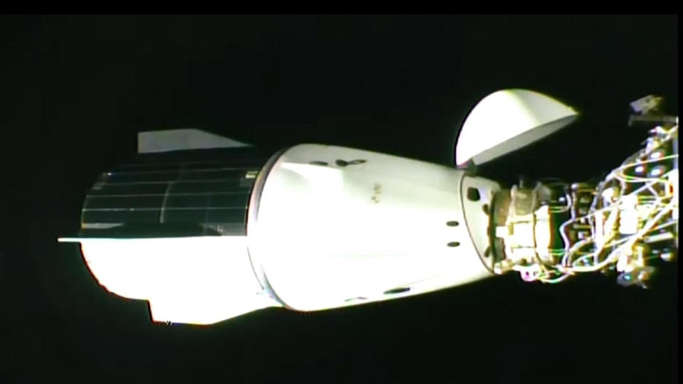 a white conical capsule is seen docked at the international space station, with the blackness of space in the background.