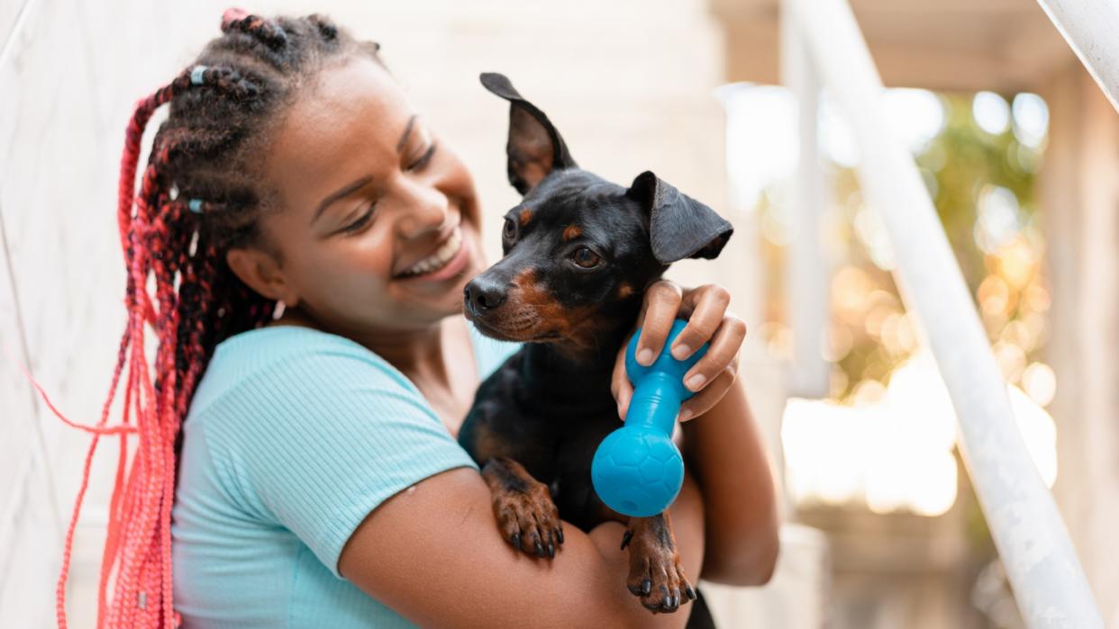 a smiling woman holds her small dog and a blue dog toy