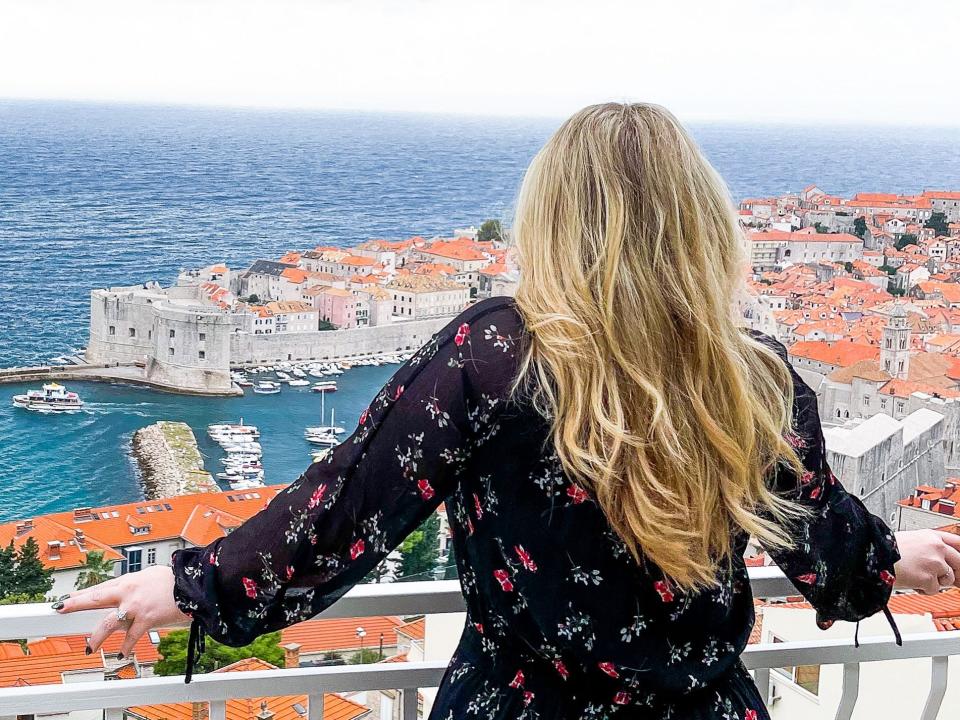A blonde woman with her back to the camera standing at a railing looking at a view of red roof buildings and the sea.