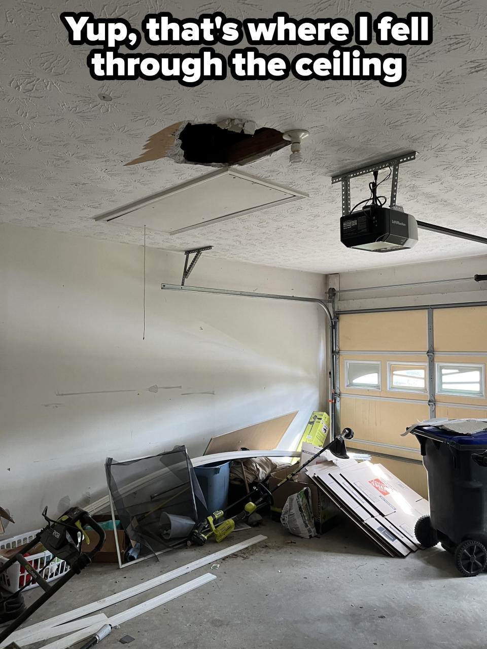 Garage with a hole in the ceiling, clutter on the floor including a trash can, boxes, and various tools. Light fixture hangs near the damaged area