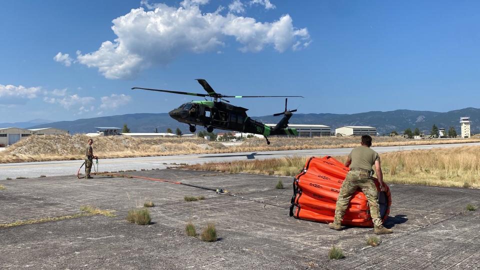 Spc. Ian Little, right, and Sgt. Ryan King, left, assigned to A. Co., 4th Battalion, 3rd Aviation Regiment, 3rd Combat Aviation Brigade, 3rd Infantry Division, prepare to hook up a Bambi bucket to a UH-60 Blackhawk during firefighting operations in Greece, Aug. 1, 2023. (Sgt. 1st Class Noel Perez/Army)