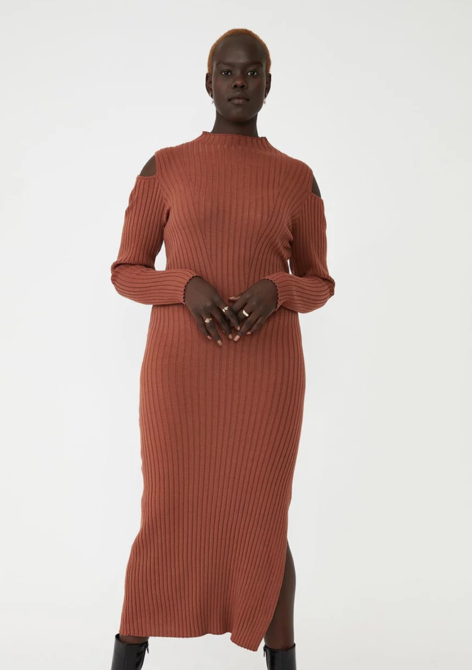 A model wears a deep ochre coloured knot dress with high neck and cutaway shoulders over short boots.