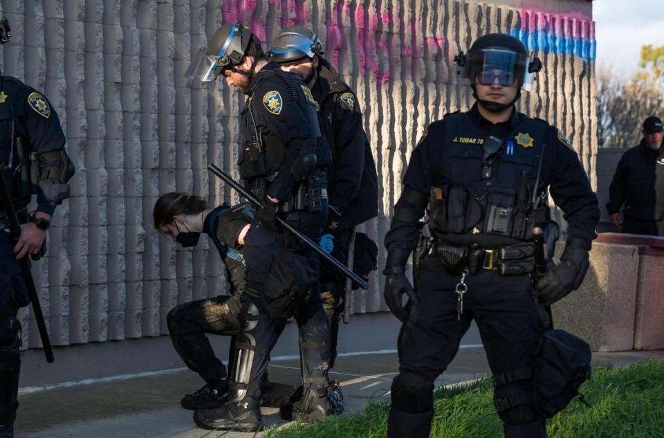 A protester is handcuffed by University of California police officers outside the U Center before an appearance by Charlie Kirk, founder of the conservative nonprofit Turning Point USA, on Tuesday at UC Davis. Behind them a section of the wall is seen spray painted in colors used to signify transgender rights.