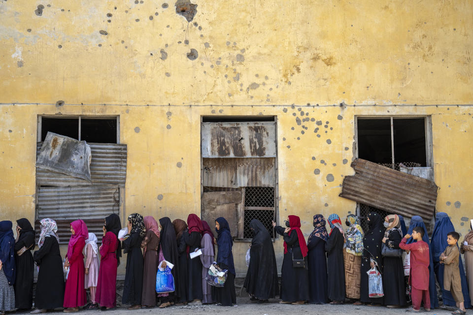 A long line of Afghan women in full-length robes and head coverings waits by a warehouse.