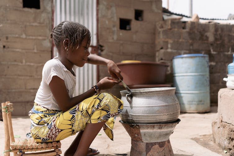 A girl sat stirring a metal pot over a charcoal stove.