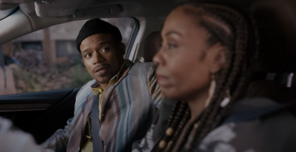 Man in a striped jacket and black beret sits in a car with a woman with braided hair and hoop earrings. Both have serious expressions. Names unknown