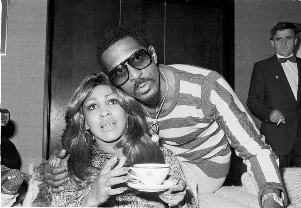 A black and white photo of Ike Turner embracing Tina Turner, who is holding a cup and saucer.