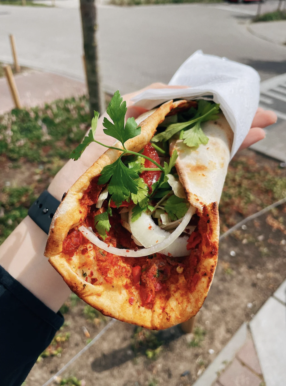 Person holding a wrapped sandwich with tomatoes, onions, and parsley