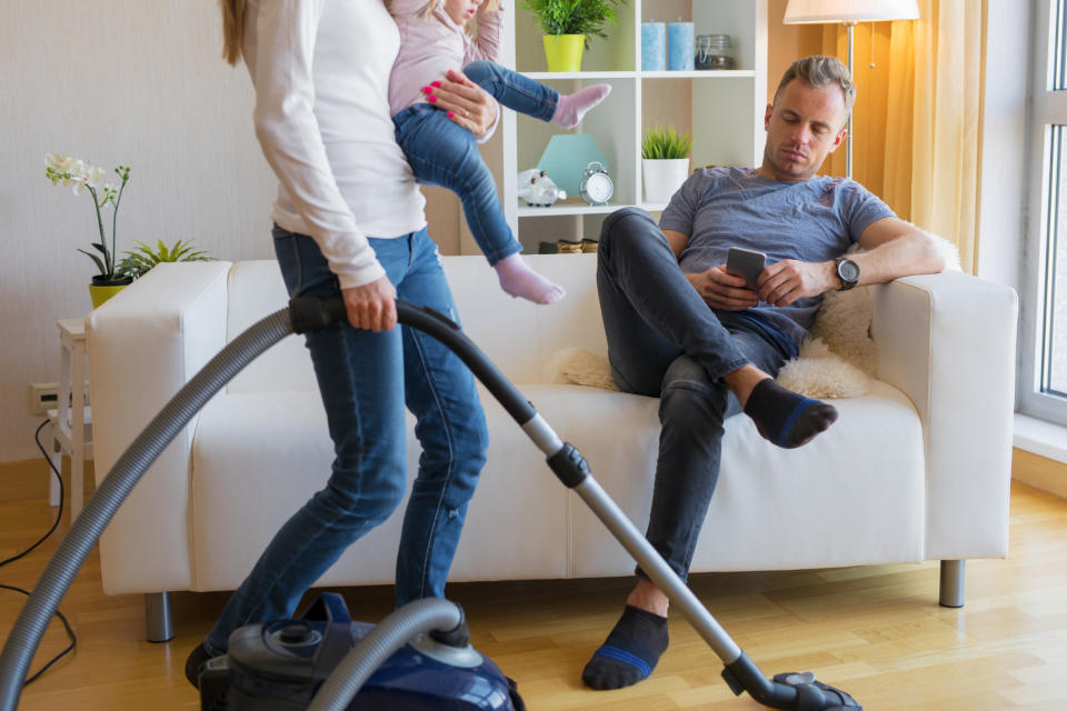 A woman vacuums while holding a child. A man sits on a couch looking at his phone