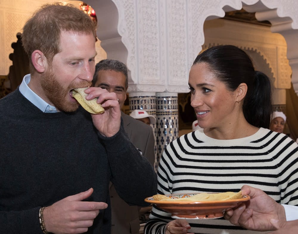 Prince Harry eating a sandwich as Meghan Markle looks on holding a plate