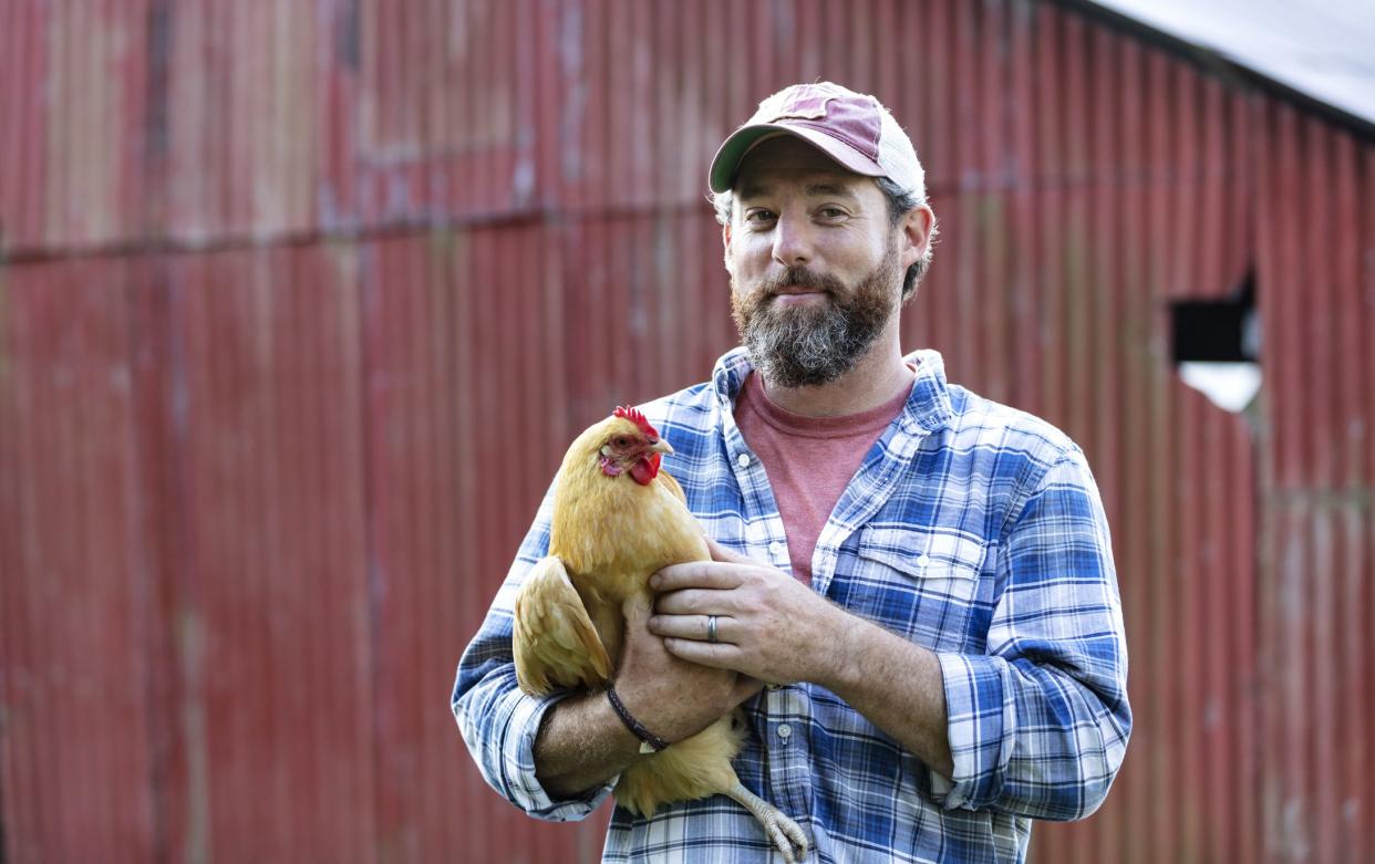 A farmer holding a chicken in his arms, smiling the camera, standing in front of a red barn. He is a mature man in his 40s with a thick beard, wearing a plaid shirt and cap.