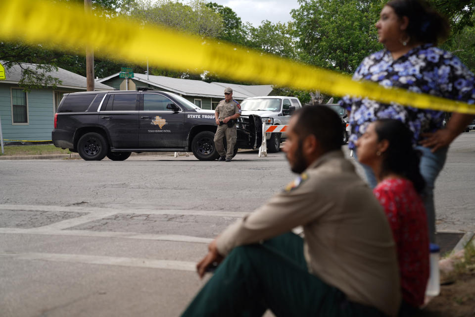 Two people sit one stands by a curb alongside yellow police tape a dozen yards from a state trooper, who is leaning against a police vehicle.