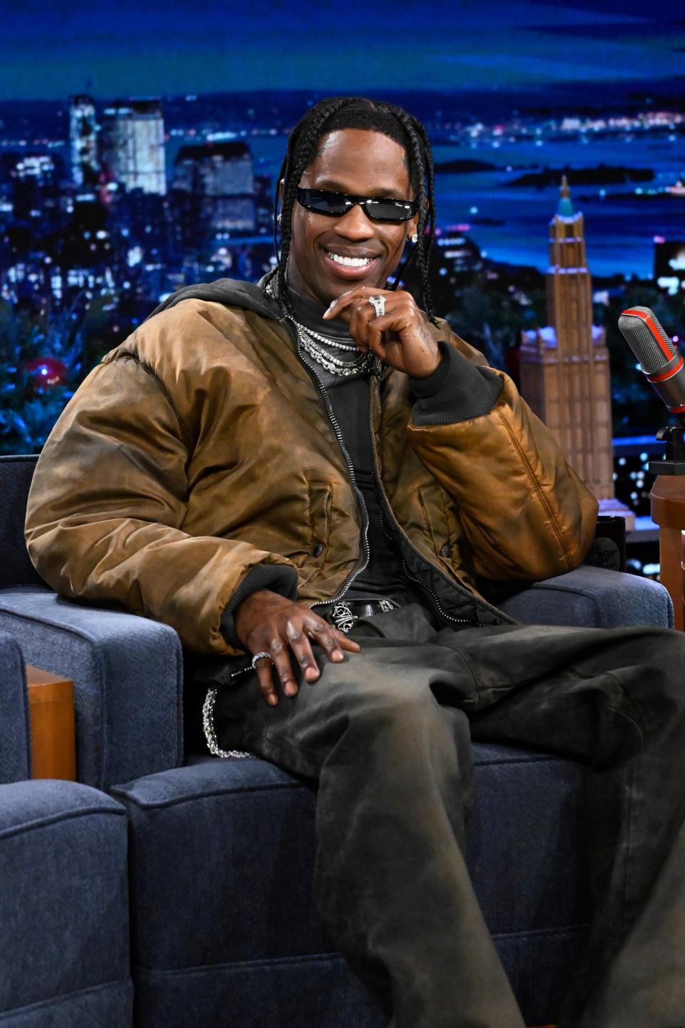 Travis Scott sits on a talk show set in a casual outfit with a large jacket, sunglasses, and jewelry, smiling with his hand partially covering his mouth