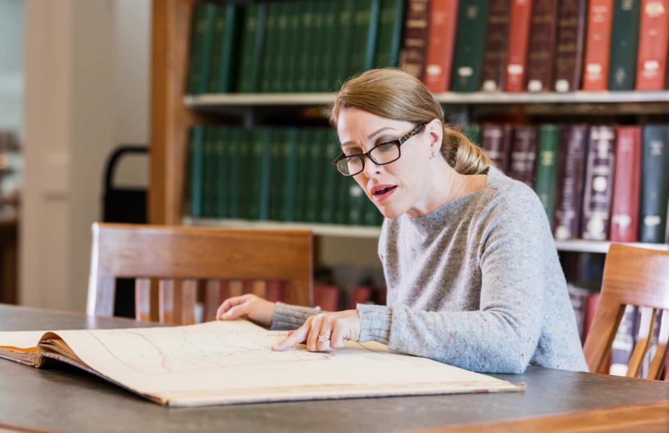 A mature woman in her 40s doing research in the library, sitting at a table looking at a large old book.