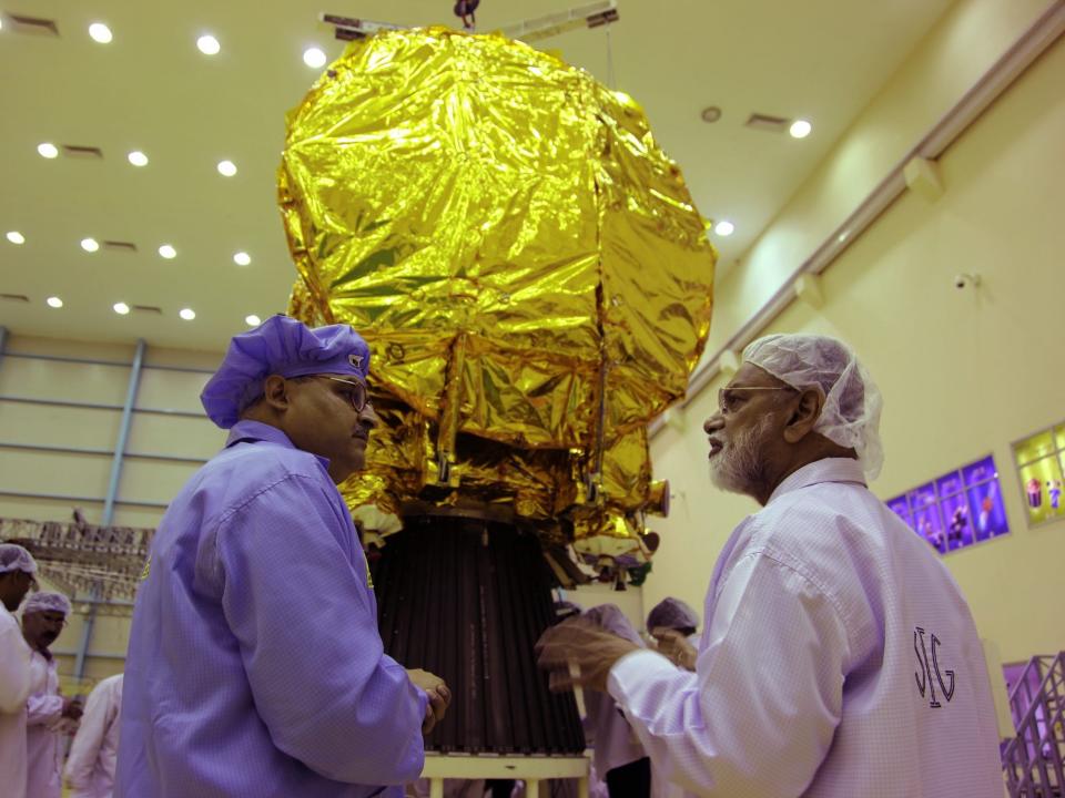 two men in harnets and clean suits talk in front of a gold foil covered spacecraft in a large cleanroom