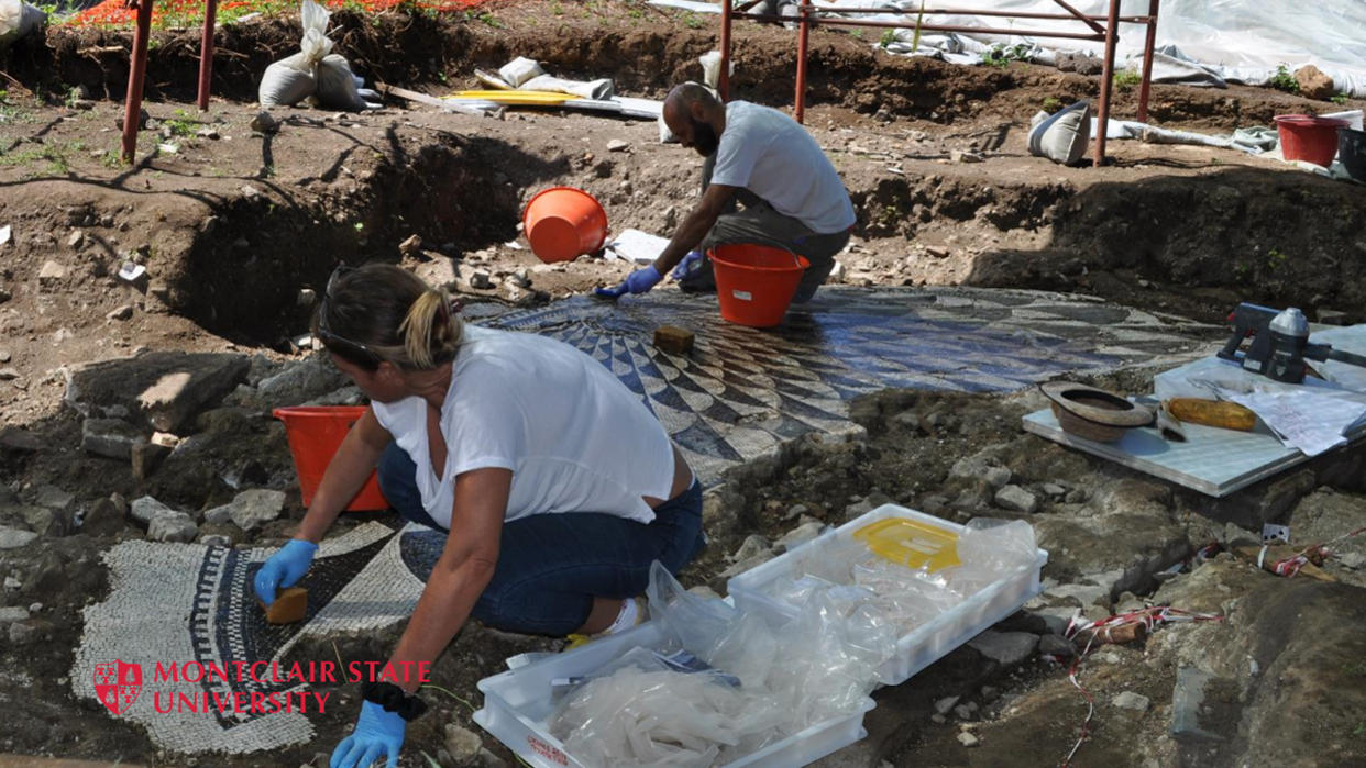  Two restorers work on their knees on a Roman-era black and white mosaic surrounded by dirt.  