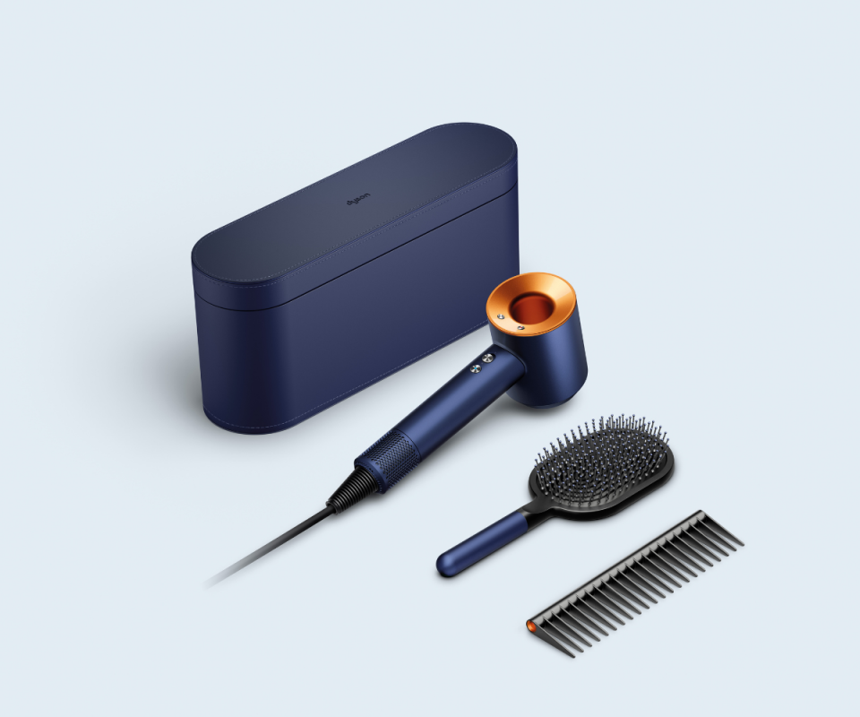 The Prussian blue dyson box next to the matching hair dryer, black brush and comb infront of a pale blue background.