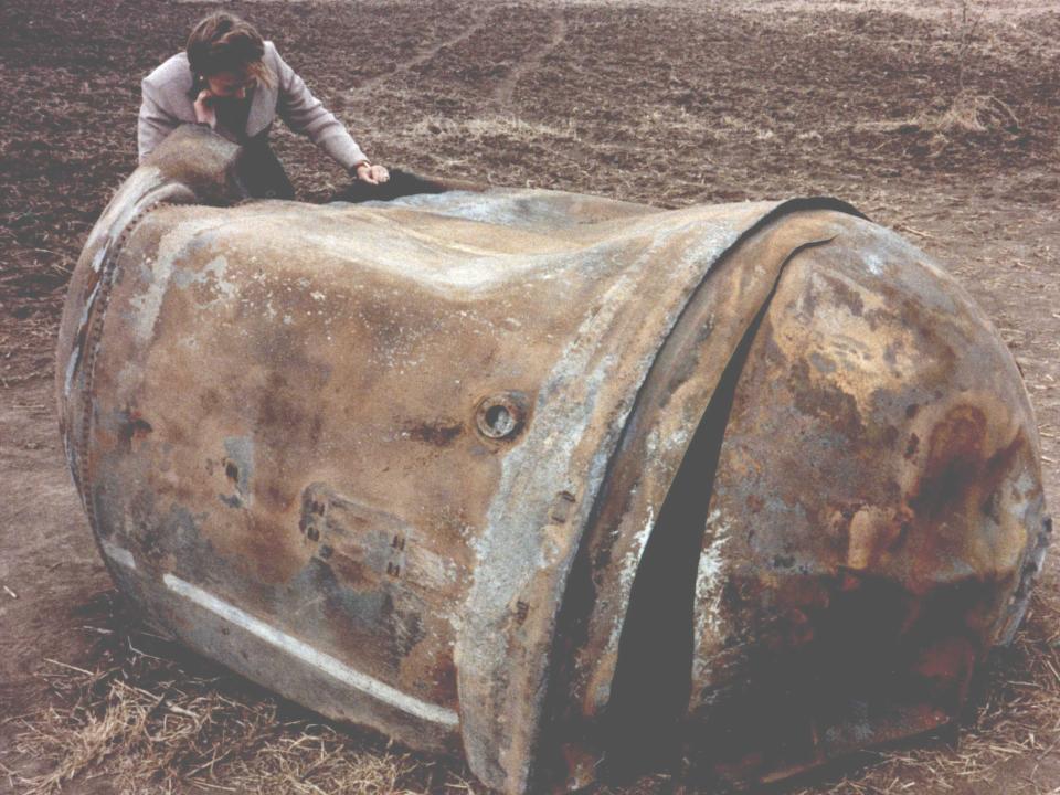 A man stands over a piece of space junk that lnded in an empty Texas field. It looks like a giant, rusted and dented trash can toppled on its side.