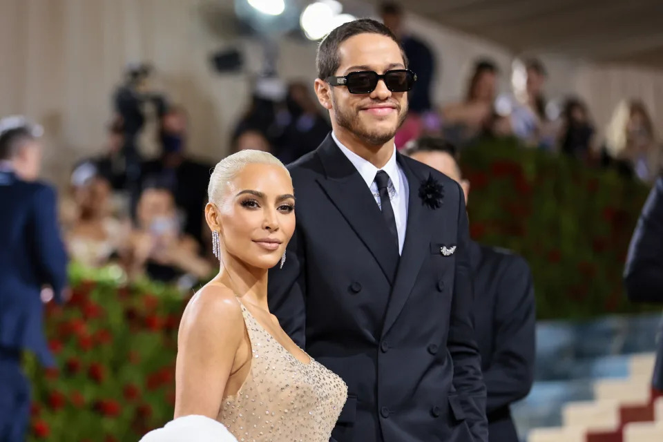 Kim Kardashian and Pete Davidson on the red carpet. Kim is wearing a glamorous, fitted gown, while Pete is dressed in a classic black suit with sunglasses