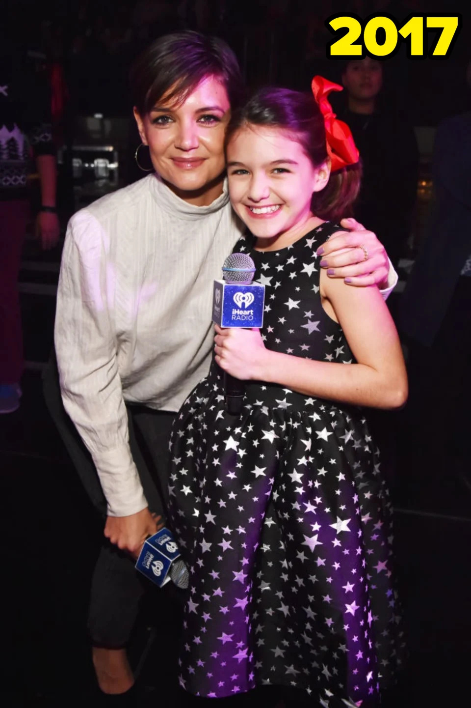 Katie Holmes and Suri Cruise smile while posing together. Suri holds a microphone and wears a star-patterned dress with a red bow in her hair