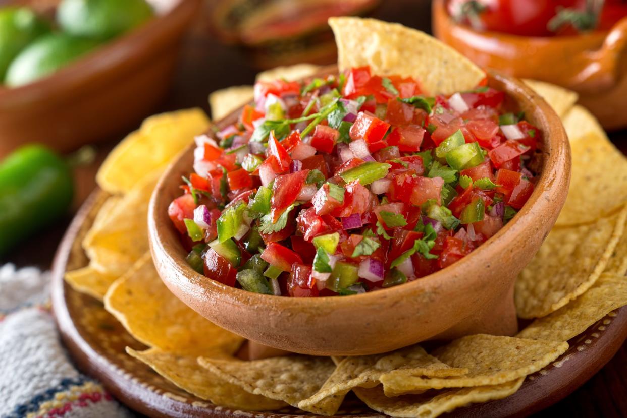 Fresh & spicy salsa in a wooden bowl surrounded by tortilla chips on a brown ceramic plate with a blurred background of ingredients