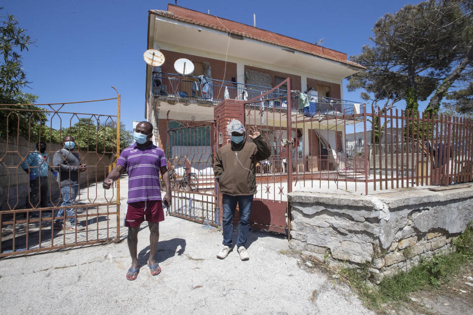 In this photo taken on Monday, April 27, 2020, Jimmy Donko, 43 from Ghana, right, waves in front of the house where he lives with other 45 men, from Nigeria and Ghana, in Castel Volturno, near Naples, Southern Italy. The home has no running water, the dilapidated electrical system doesn't reach many rooms that are in the dark. They are known as “the invisibles,” the undocumented African migrants who, even before the coronavirus outbreak plunged Italy into crisis, barely scraped by as day laborers, prostitutes and seasonal farm hands. Locked down for two months in their overcrowded apartments, their hand-to-mouth existence has grown even more precarious with no work, no food and no hope. (AP Photo/Alessandra Tarantino)