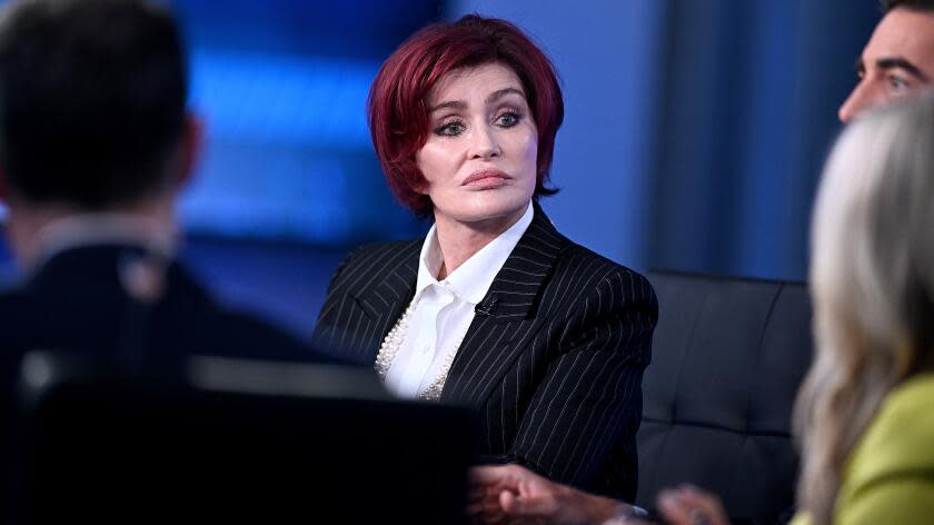 Sharon Osbourne is sitting on a panel, listening with a serious face, wearing a black pinstripe suit and white shirt