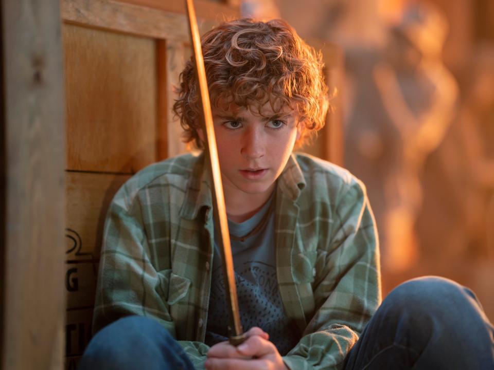 percy jackson in the disney plus live action show, crouching behind a crate and intently holding a gleaming golden sword