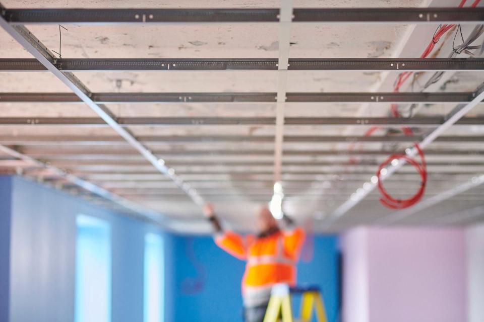 A worker in an orange vest tends to a ceiling in a blurry background. 