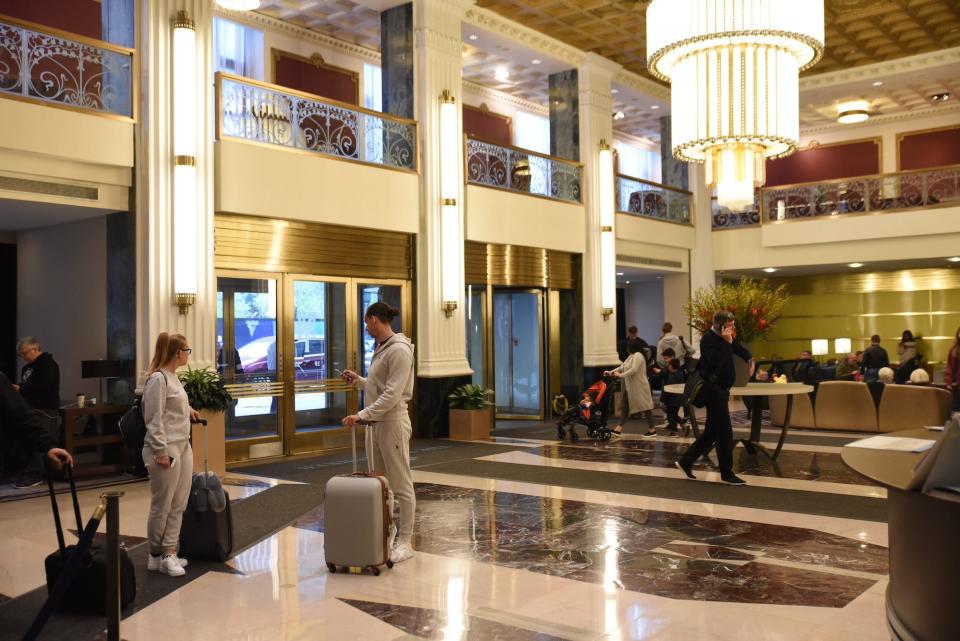 The lobby of the New Yorker Hotel, which has gilded accents, floors that look like marble, and Art Deco elements including a chandelier and an upper balcony level.