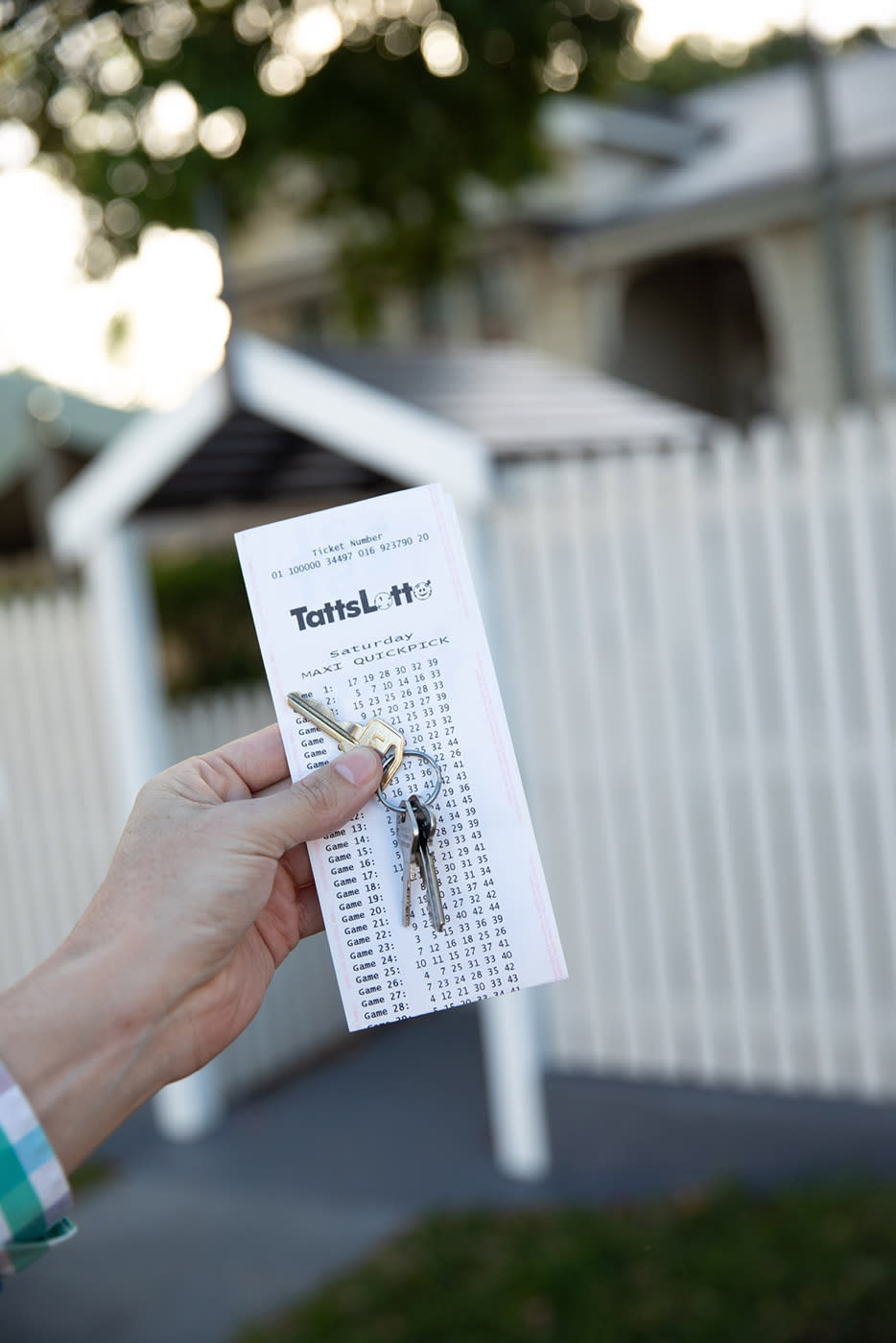 A hand holds a set of house keys and a TattsLotto ticket in front of a white picket fence.