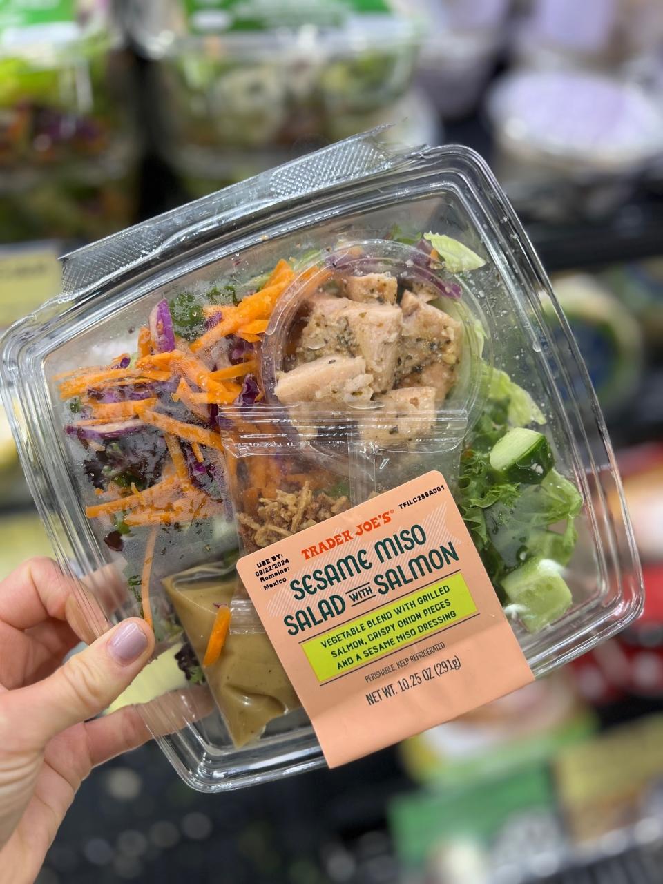 A hand holds a Trader Joe's Sesame Miso Salad with Salmon container featuring vegetable blend, grilled salmon, and a sesame miso ginger dressing