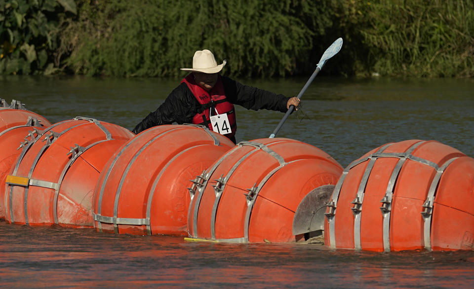 A kayaker wearing a cowboy hat floats behind large orange buoys.