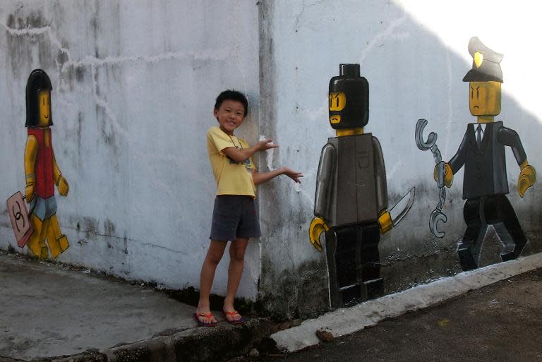 A boy poses for pictures with a mural by Lithuanian artist Ernest Zacharevic, which shows a Lego woman with a Chanel bag (L), a Lego robber armed with a knife (2nd, R) and a Malaysian authority (R) in Johor Bahru on November 13, 2013