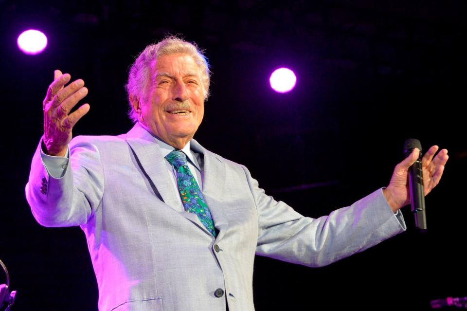 Tony Bennett in a waist-up color frame, smiling and gesturing wide with both arms.