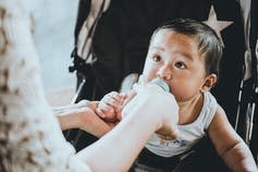 A baby looks at his mother while sucking on his bottle in a stroller.