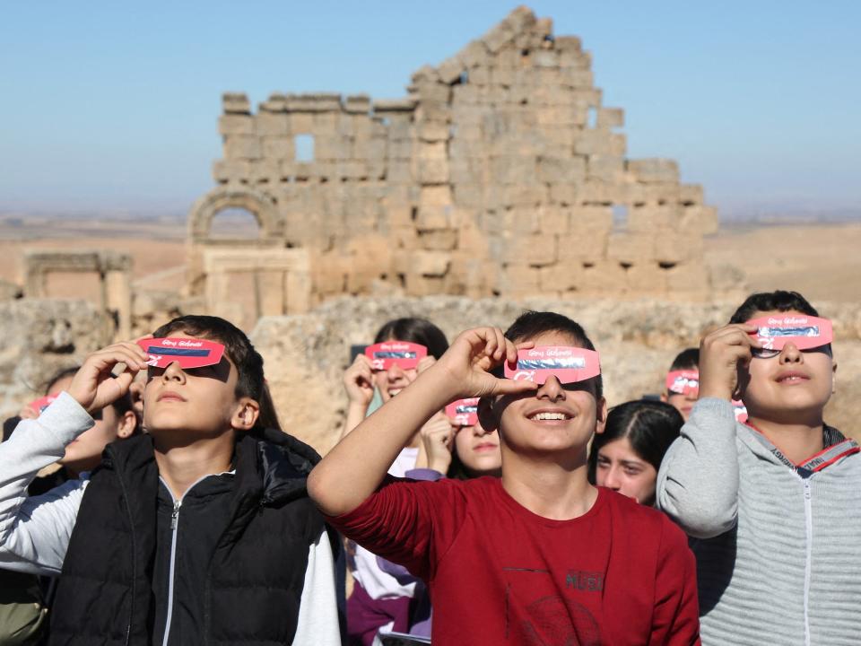 huddle of people look up toward the sun while holding pink eclipse glasses up to their eyes in front of a wall of sand colored bricks in desert landscape