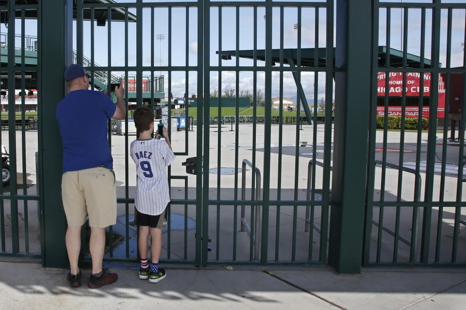FILE - Cubs fans take photos through the locked gates at Sloan Park, the spring training site of the Chicago Cubs, in Mesa, Ariz., after Major League Baseball suspended the rest of its spring training game schedule because of the coronavirus outbreak, Friday, March 13, 2020. Spring training games might not count in the official standings, but they certainly count for the pocketbooks of business owners in Arizona and Florida. They're also a much-anticipated destination for fans who come for the warm sunshine and the laid-back atmosphere. (AP Photo/Sue Ogrocki, File)