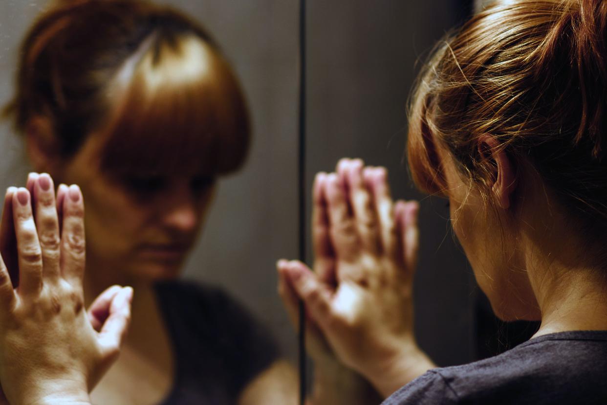 Distraught woman with both her hands on the mirror, she is facing the mirror and looking down, her face is reflected in the mirror on the left