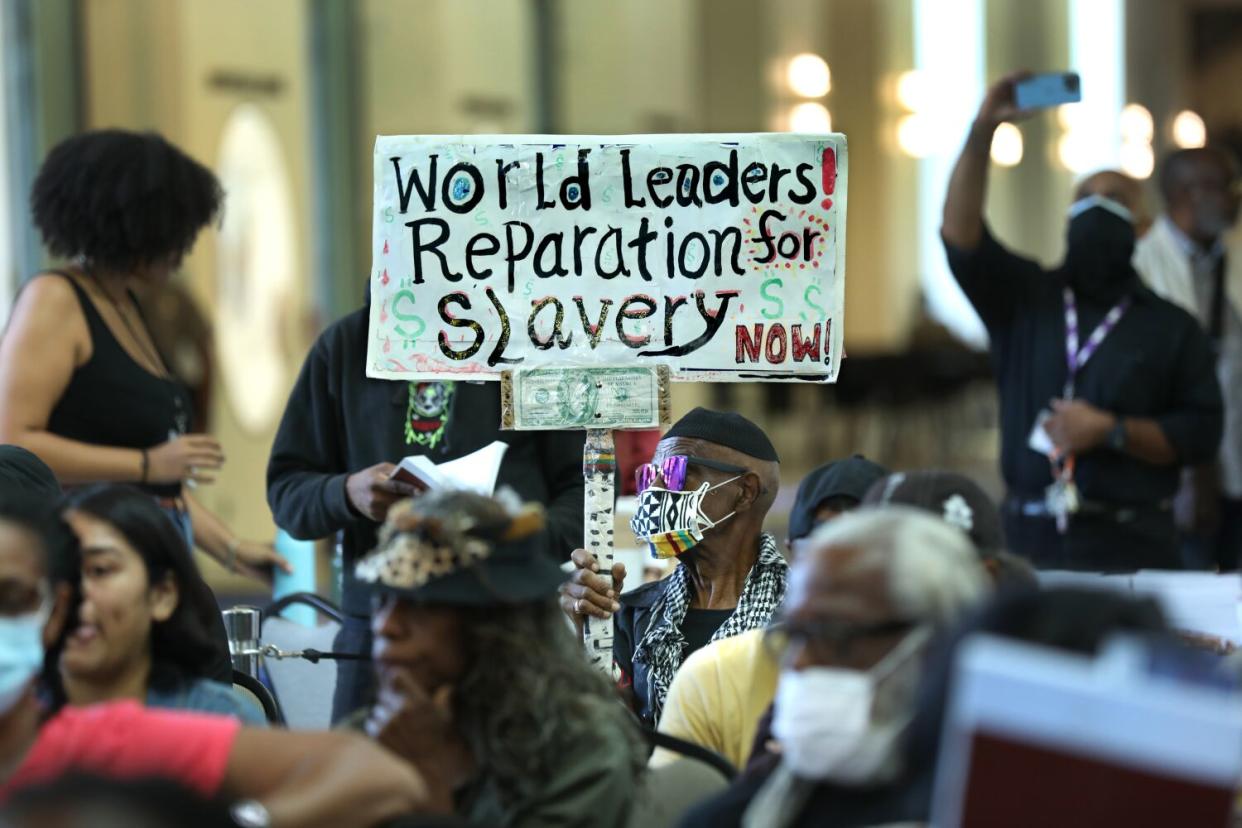 Members of the public at a meeting, one holding a sign reading, "World leaders! Reparations for slavery now!"