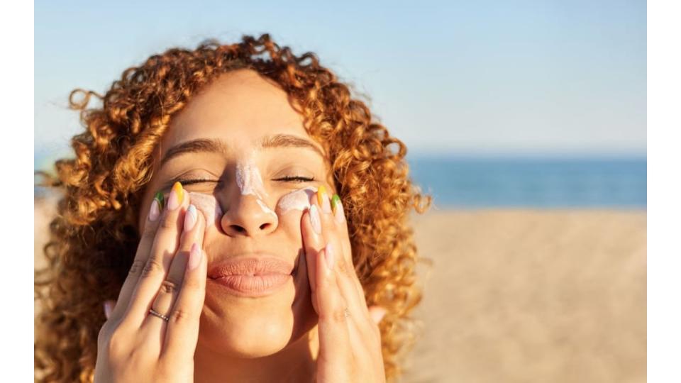 Young woman applying sunscreen to her face on the beach in a summer sunset.